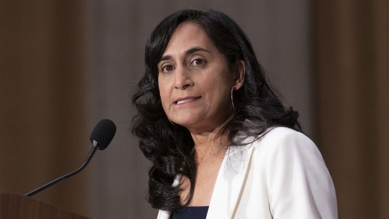 A woman with dark hair wearing a white blazer stands at a podium with a mic