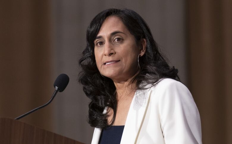 A woman with dark hair wearing a white blazer stands at a podium with a mic