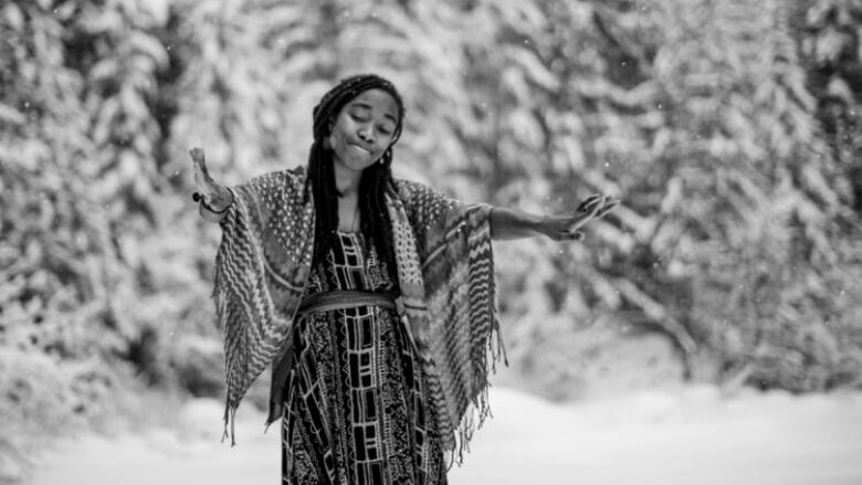 A woman with black hair wearing a shawl is dancing in a snowy field. In the background, there are trees covered in snow. 