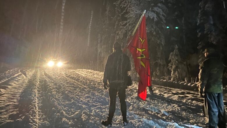 Headlights from a car makes silhouette of a person holding a red flag in the snow.