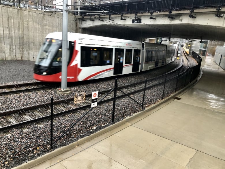 A red-and-white train travels on tracks under a concrete bridge.