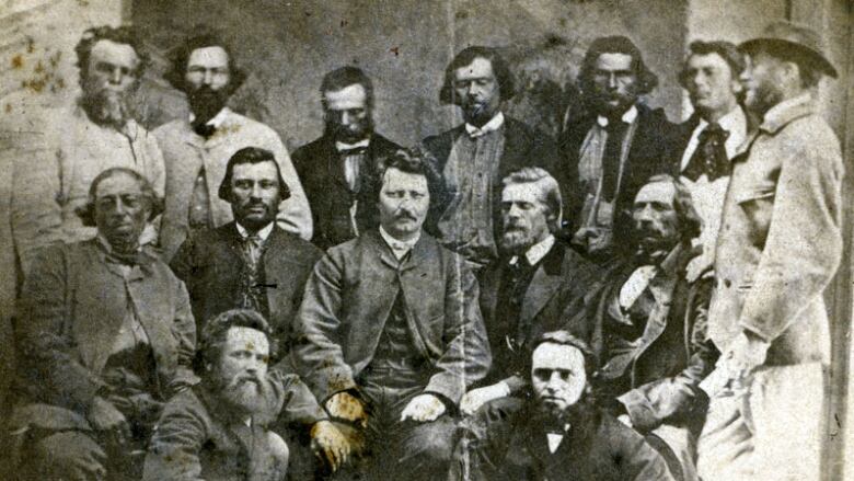 Black and white photo of a group of 15 men from the 1870s. Most are standing, surrounding one man seated in the centre.