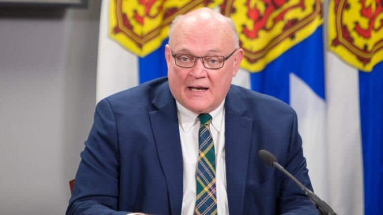 A man is shown speaking in front of a Nova Scotia flag.