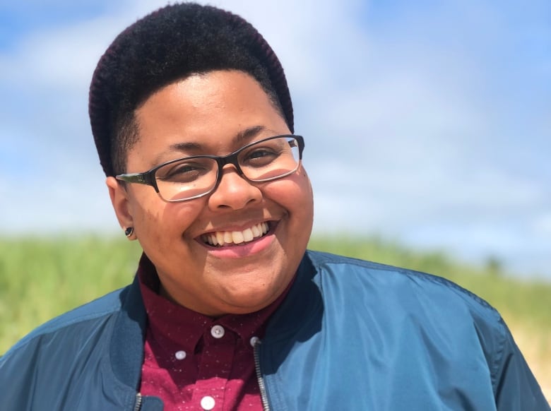 A Black woman smiling wearing glasses and a wine shirt and blue jacket.