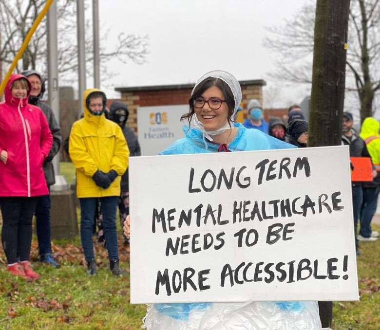 A woman in a wedding dress and a poncho standing in the rain holding a sign that says 