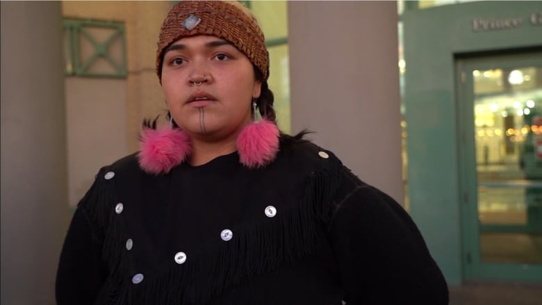 A photo of a woman with pink fur pom-pom earings leaves custody. She has a septum piercing with a smal silver ring and a tradition face tattoo with two lines down her chin. 