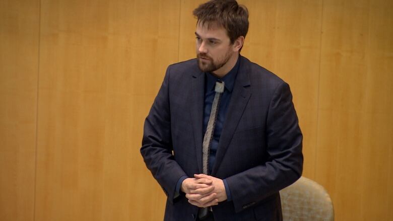 A white man with brown hair, facial hair and wearing a suit is standing in a room with his hands clasped together.