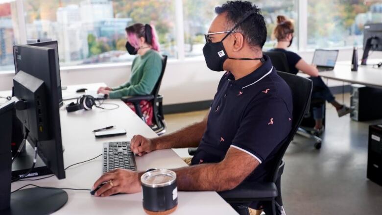 Individuals wear masks while working at computers in an office. 