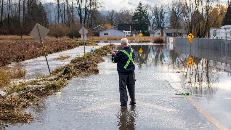 A person wearing high-vis clothes stands in ankle-deep floodwaters on a road and points their phone at signs on the road.