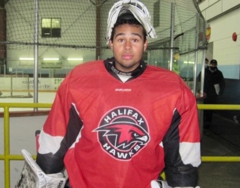 A Black teen is wearing hockey gear with a shirt that reads 'Halifax Hawks