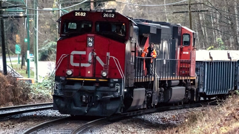 A red and black CN train engine is seen on a railway, with an engineer standing on the train engine's platform. 