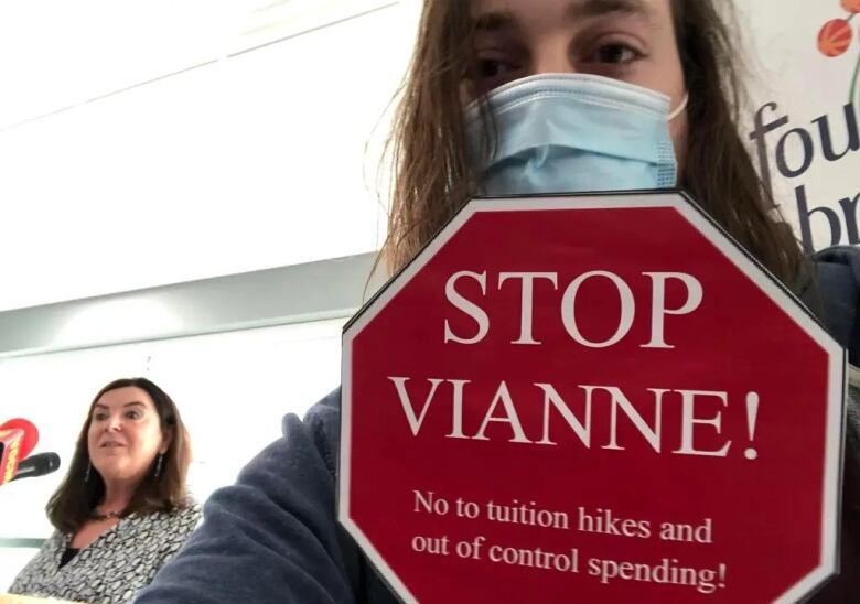 A man with long hair, wearing a non-medical mask, holds a stop sign that says 