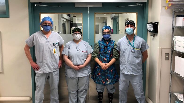 Four medical workers in scrubs and surgical masks stand in front of a hospital operating room.