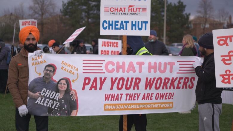 People standing with banners and placards saying pay your workers what they're owed. 