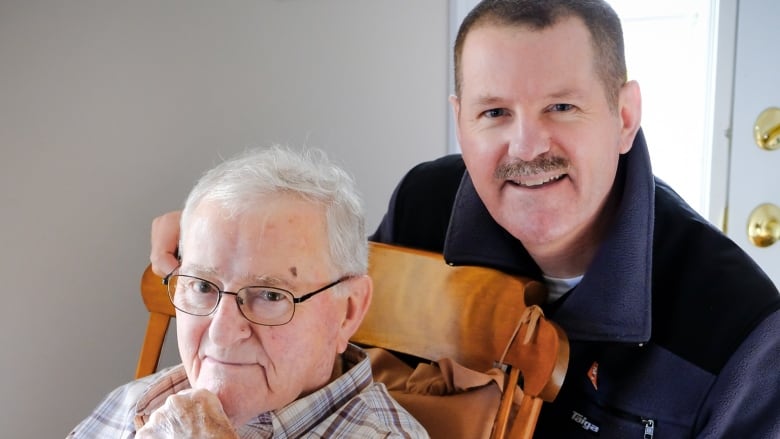 An older man sits in a rocking chair while a younger man stands behind him. 