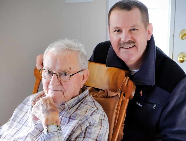 An older man sits in a rocking chair while a younger man stands behind him. 