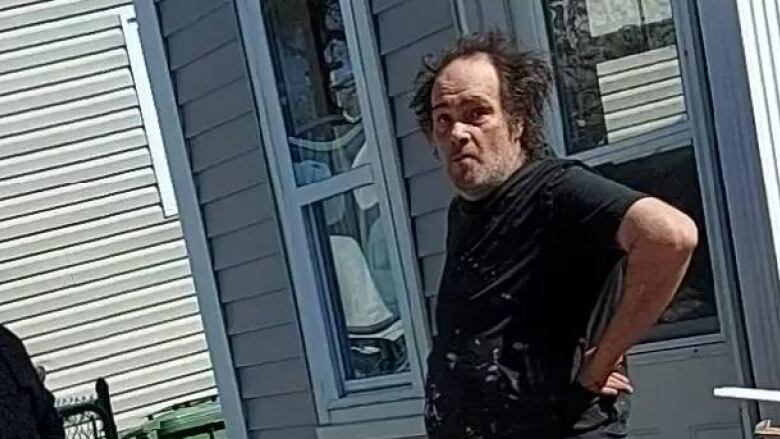 A man with a u-shaped hairline and disheveled hair is wearing a paint-splattered black t-shirt. His left arm is on his hip.