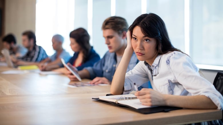 A woman holds a pen and slouches over a planner in a meeting while looking bored.