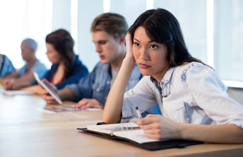 A woman holds a pen and slouches over a planner in a meeting while looking bored.