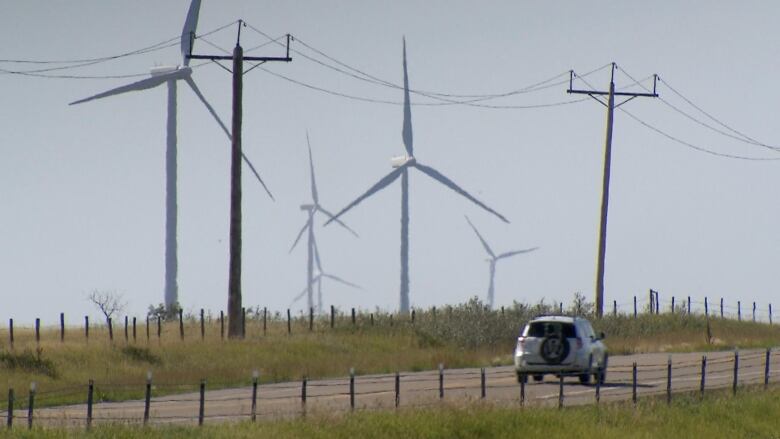 Wind turbines are seen behind electricity transmission lines along a rural road.