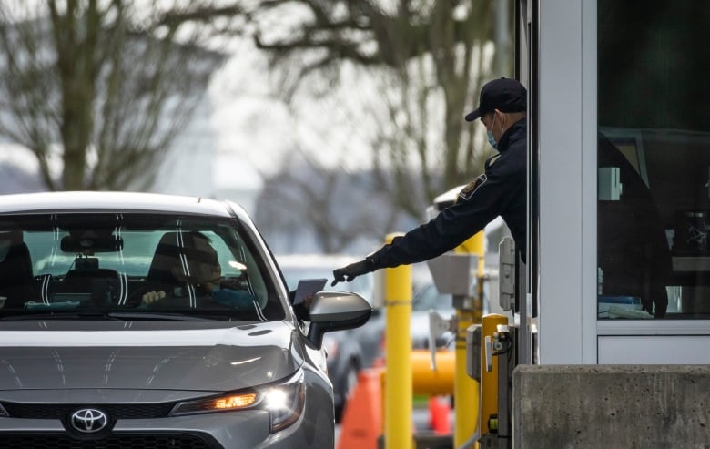 An officer at a border crossing reaches out to get someone's documents from a car window.