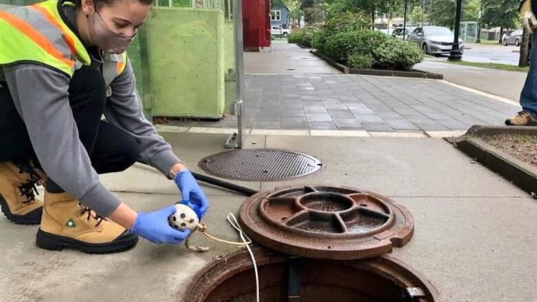 A person with a mask stands by a sewer in the street and holds a device. 