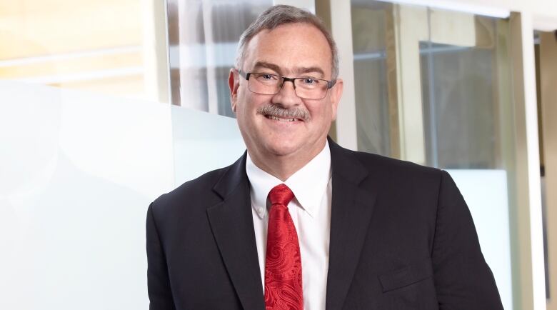 Dr. Jon Meddings wears a black suit and red tie and leans with his right shoulder against a window.