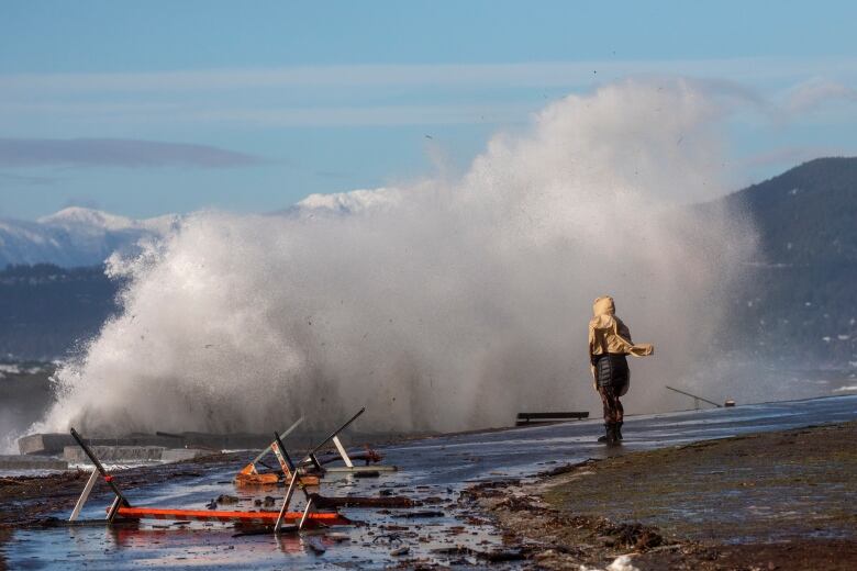 A woman in a rain jacket stands on a sidewalk with a huge wave crashing in front and over her. On the side are fallen over construction warning signs. 