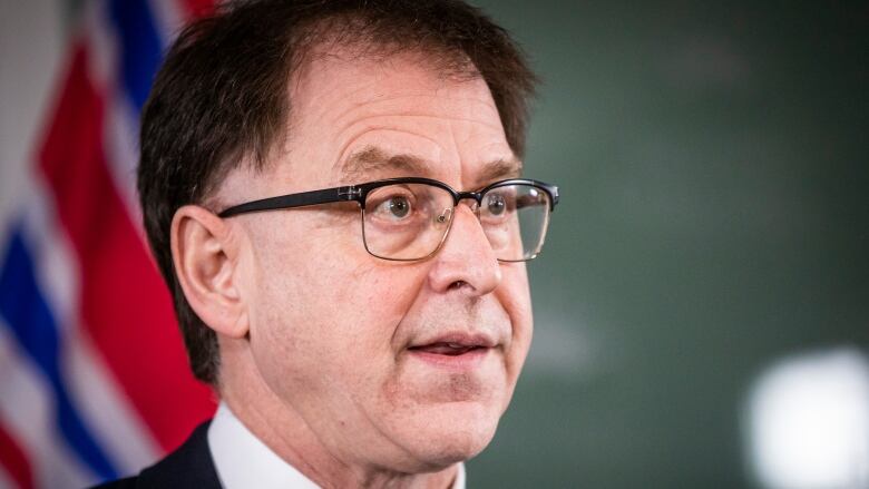 A close-up shot of a man in glasses in mid speech with a B.C. flag in the blurred out background.