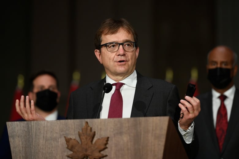 A man in a black suit stands at a podium in a dark room.