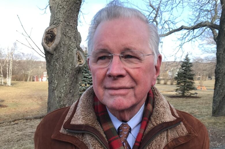 Older man with white hair and glasses, standing next to a tree.