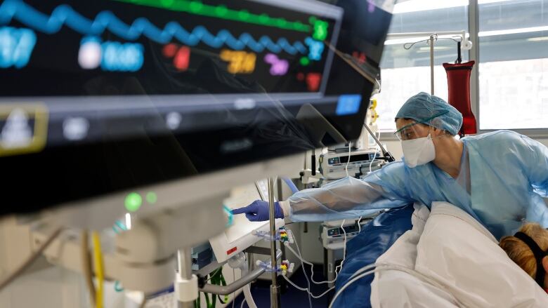 A nurse reaches toward a piece of medical equipment beyond the top of a patient's bed, while a monitor is seen in the foreground.