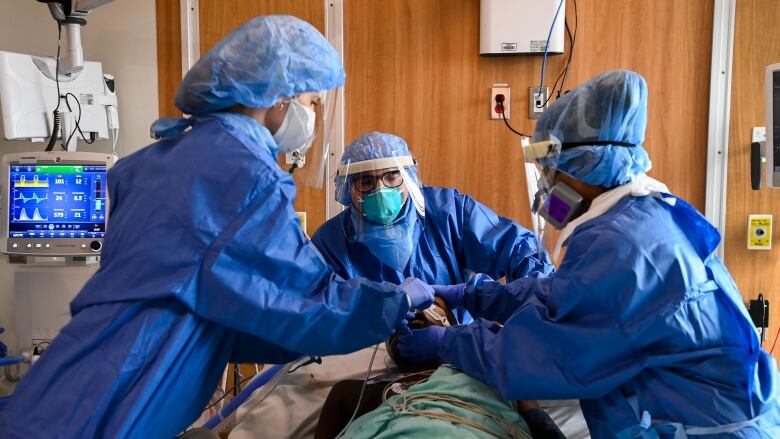 Three hospital staff members in blue gowns and facemasks work on a patient lying on a stretcher.