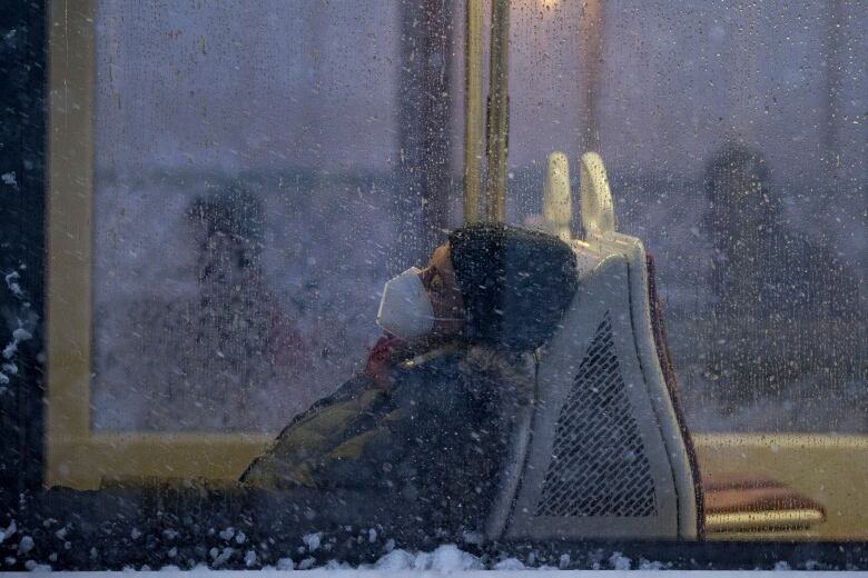 A person wearing a mask and a winter coat and hat sits on public transit with their eyes closed