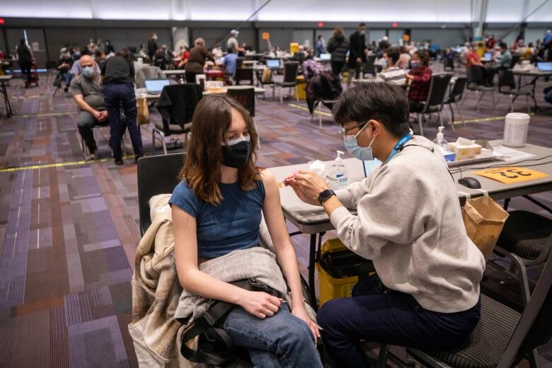 A girl receives a vaccine in a convention hall. Behind her, dozens more people are seen getting vaccinated.