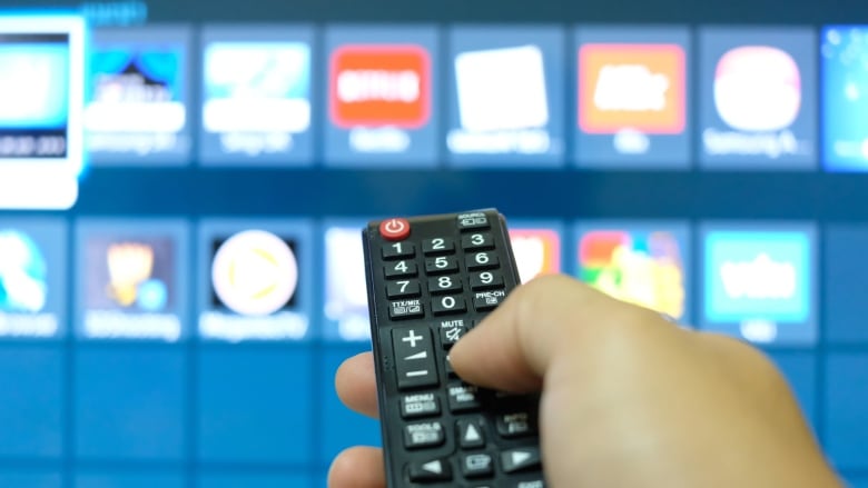 A hand holding a remote is seen in front of a TV screen showing out-of-focus apps.