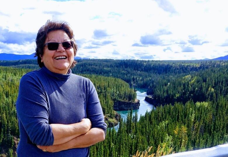 A smiling woman stands poses before a scenic vista of forested hills and a river.