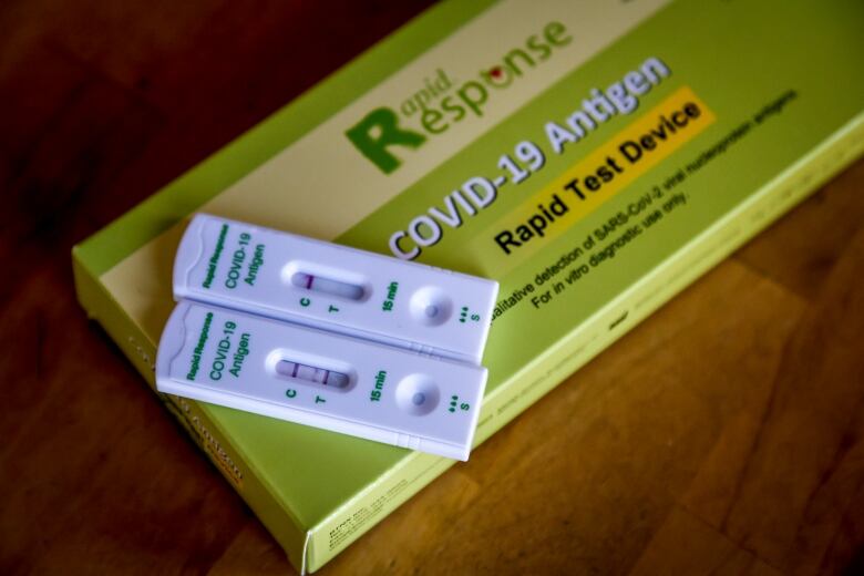 Two COVID rapid tests sit on top of a green box, marked 'Rapid Response, COVID-19 Antigen Rapid Test Device.' One has two lines -- meaning it is positive. The other has one line, meaning it is negative.