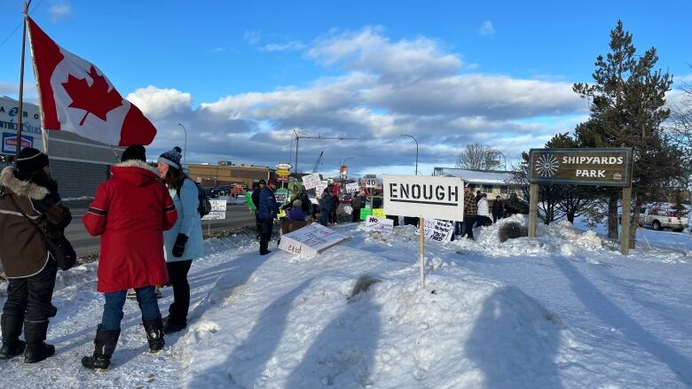 A group of people with signs and Canadian flags stands on a snowy sidewalk. 