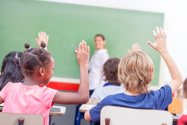 Students seated at desks raise their hands as a teacher stands at a chalkboard at the front of the classroom.