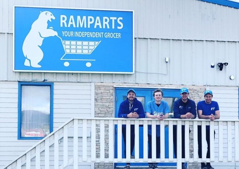 Four men stand in a row leaning on a balustrade outside a grocery store.