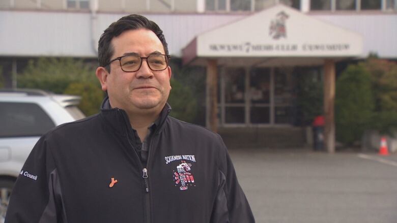 An Indigenous man wearing a black jacket that reads 'Squamish Nation' speaks to a camera outside a building.