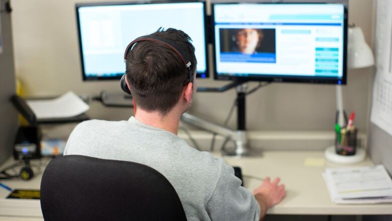 A man wearing a headset sits at a desk facing a computer screen.