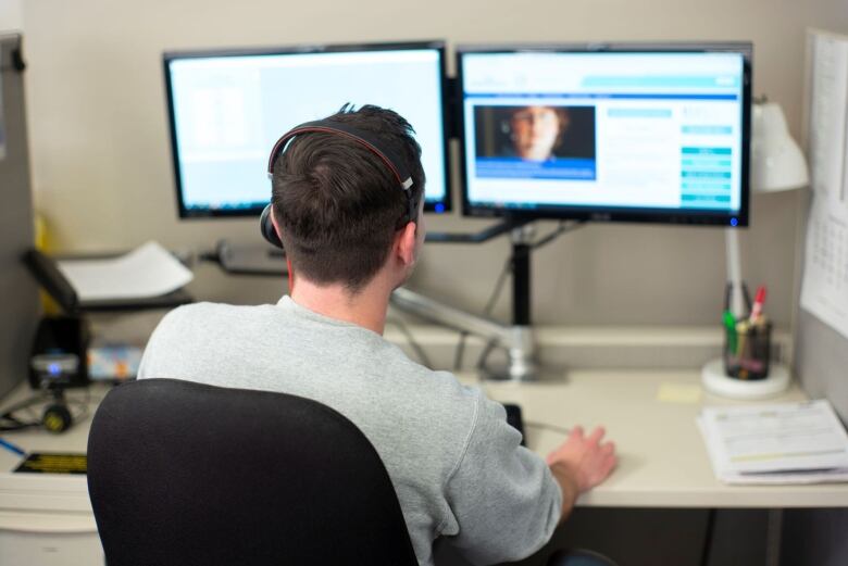 A man wearing a headset sits at a desk facing a computer screen.