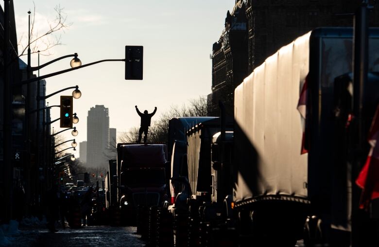 A man with his hands in the air is seen in silhouette while standing on top of one of several transport trucks parked on a downtown street.