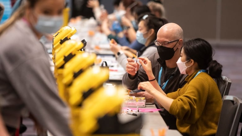 People wearing masks sit at a long table, hold syringes.