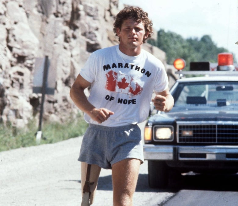 Terry Fox runs along a road with a car behind him. He is wearing a shirt that reads 'Marathon of Hope' and has a prosthetic leg.