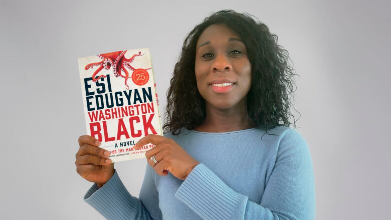 A Black woman with dark, curly hair wearing a blue sweater. She is holding a copy of the novel 