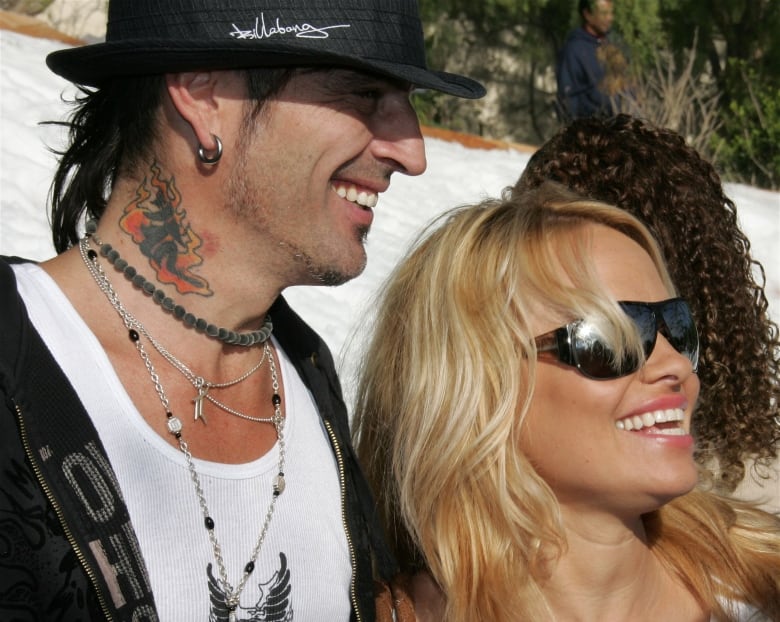 A man wearing a black hat and white T-shirt, and a woman with long blonde hair and sunglasses smile into the sunshine.