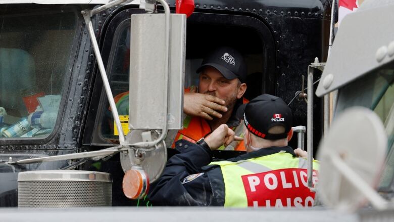 A police officer with a red 'POLICE LIASON' label on his back talks to someone in a truck in a city in winter.